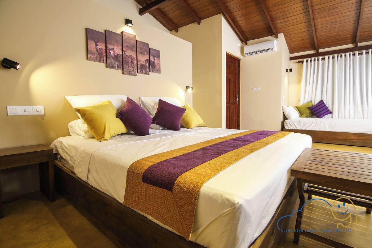A hotel room with a large bed, a wooden bench, a bedside table, and a wall-mounted air conditioner.