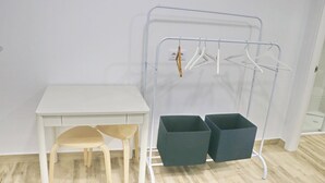 Una habitación con un escritorio blanco, dos sillas, un tendedero metálico con perchas y dos cajas de almacenamiento.