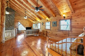 A log cabin interior with wooden floors, a stone fireplace, a bed with a wooden headboard, and a ceiling fan.