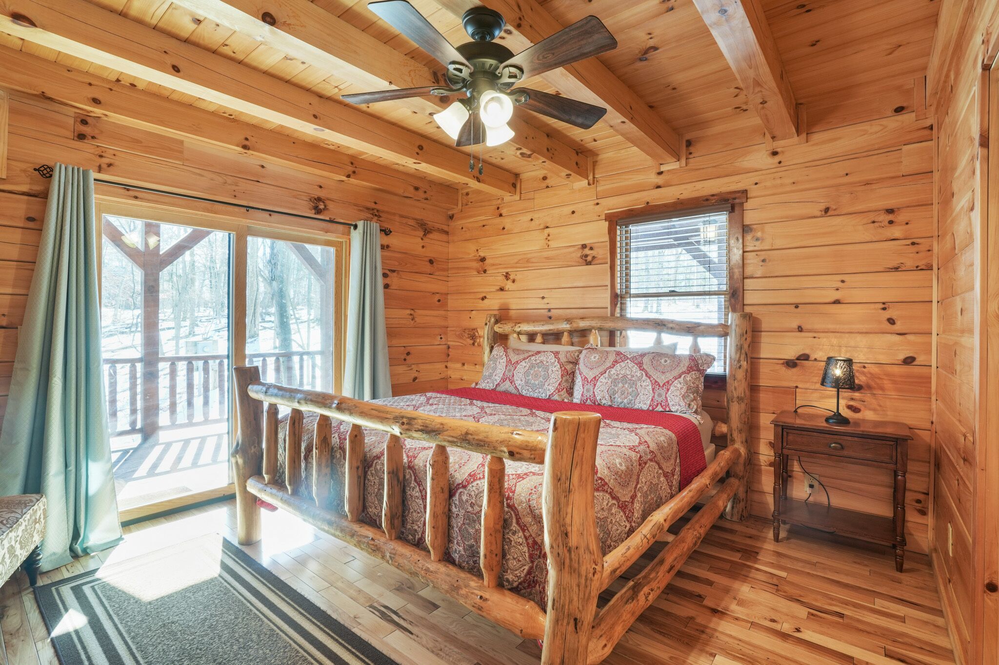 A wooden cabin bedroom with a bed, a nightstand, a ceiling fan, and large windows.