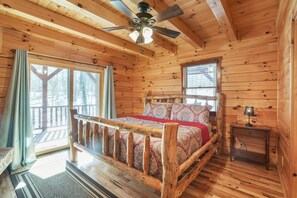 A wooden cabin bedroom with a bed, a nightstand, a ceiling fan, and large windows.