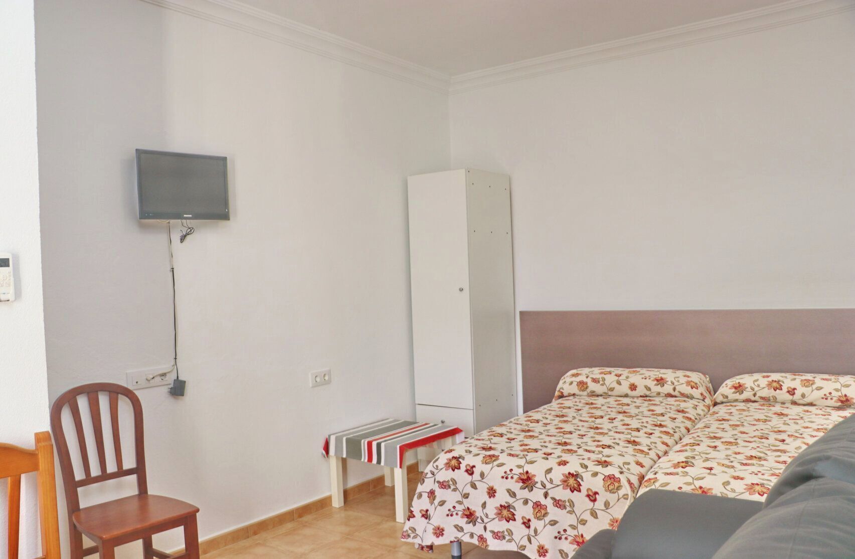 A small, simple room with a bed, a chair, a TV mounted on the wall, and a wardrobe.