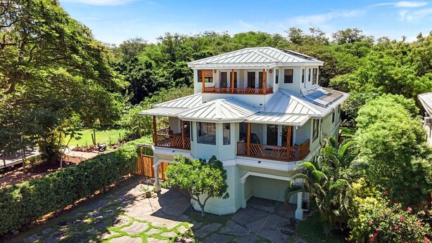 A two-story house with a balcony, surrounded by lush greenery and a stone pathway.