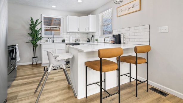 A modern kitchen with white cabinetry, a central island with bar stools, pendant lighting, and a 'be our guest' sign. - Game Room Retreat with Firepit + Gym + Sleeps 12 (Indianapolis)