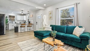 A modern living room with a teal sofa, wooden coffee table, and a kitchen in the background. - Game Room Retreat with Firepit + Gym + Sleeps 12 (Indianapolis)