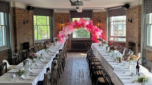 A long banquet hall with white tables, chairs, and a pink balloon arch. - The Brookmill (London)