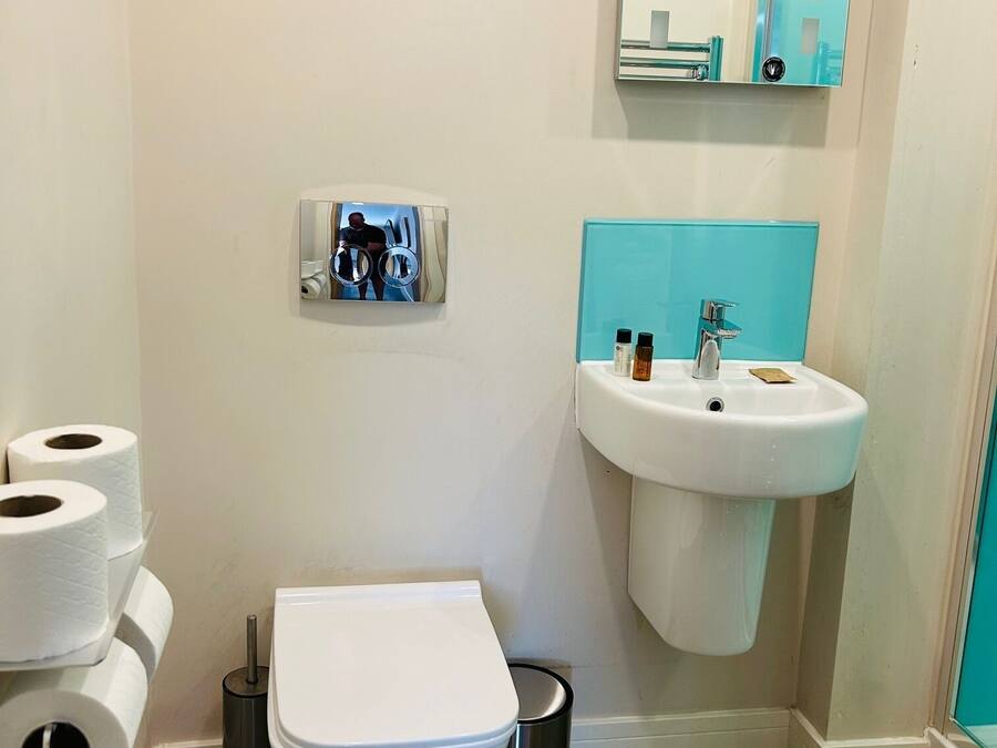 A small bathroom with a toilet, a sink, and a mirror. There are toilet paper rolls and a trash can.