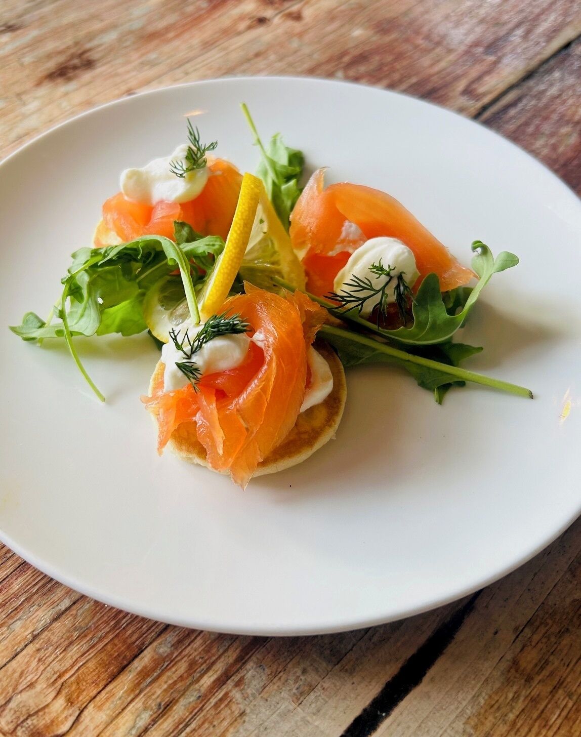 A white plate with a food arrangement featuring smoked salmon, greens, and a lemon slice.