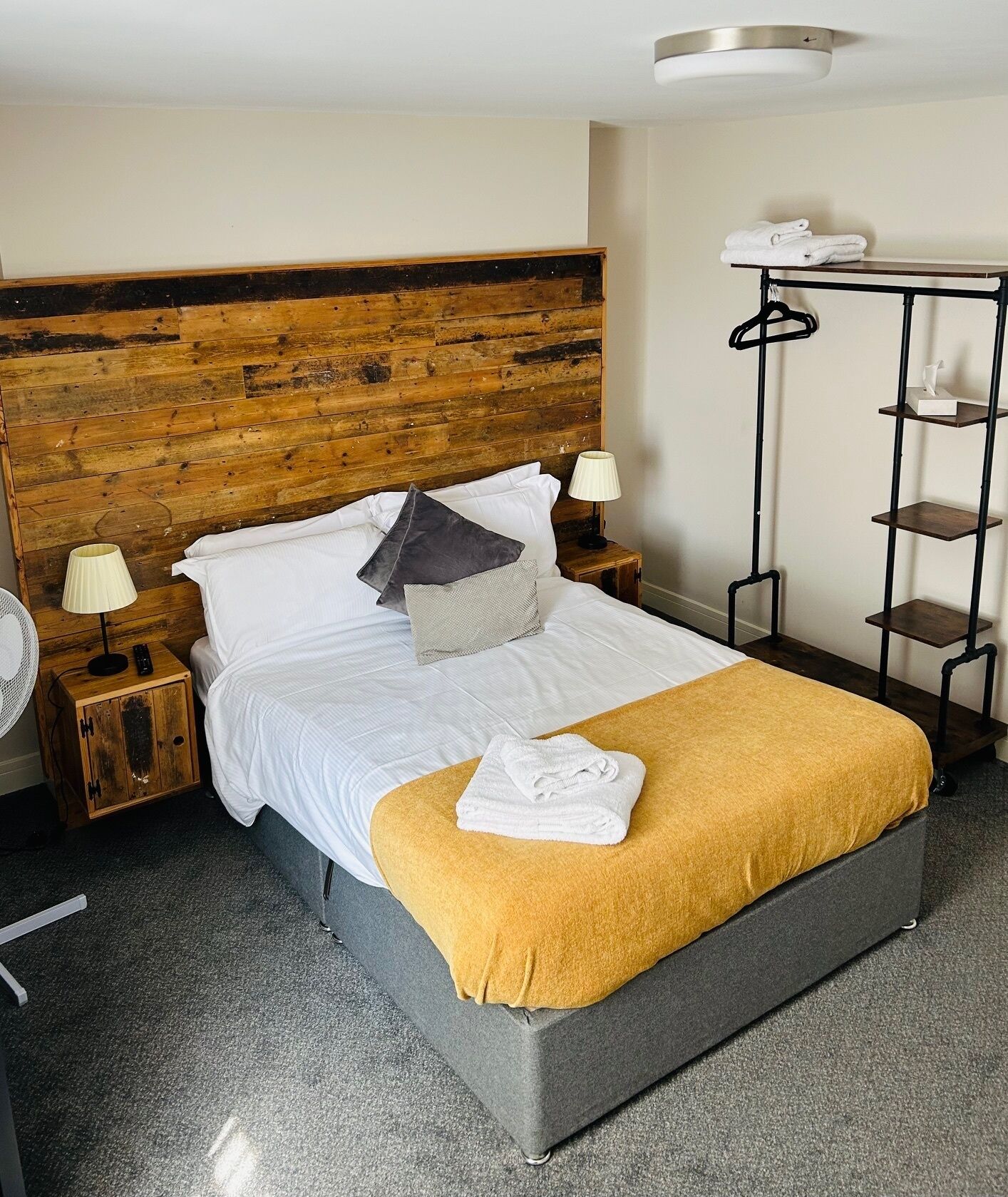 Ein ordentlich bezogenes Bett mit Holz Kopfteil, ein Nachttisch mit Lampe, ein kleiner Holzschrank und ein hoher, schwarzer Metallregal mit Handtüchern.