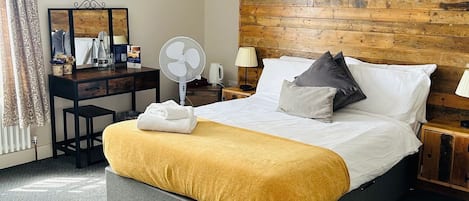 A modern hotel room with a large bed, a wooden headboard, a desk with a mirror, a fan, and a lamp.