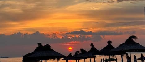 Une plage avec des parasols en chaume et des chaises longues blanches, un coucher de soleil en arrière-plan et une mer calme.