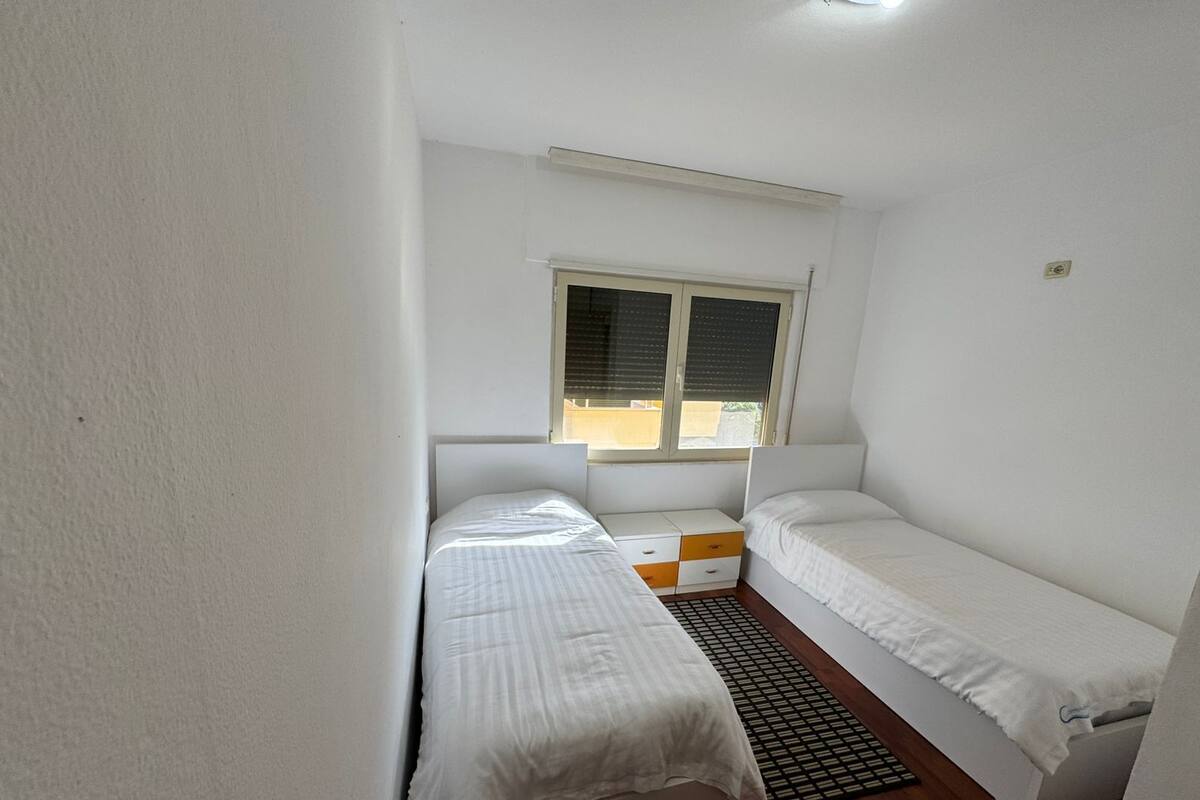 Un alojamiento pequeño y de una sola habitación con dos camas individuales, una ventana con persianas, una mesita y un radiador.
