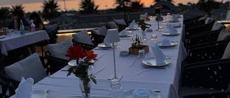 A sunset view from a restaurant terrace with tables set for dining, a hanging lamp, and a vase with red flowers.