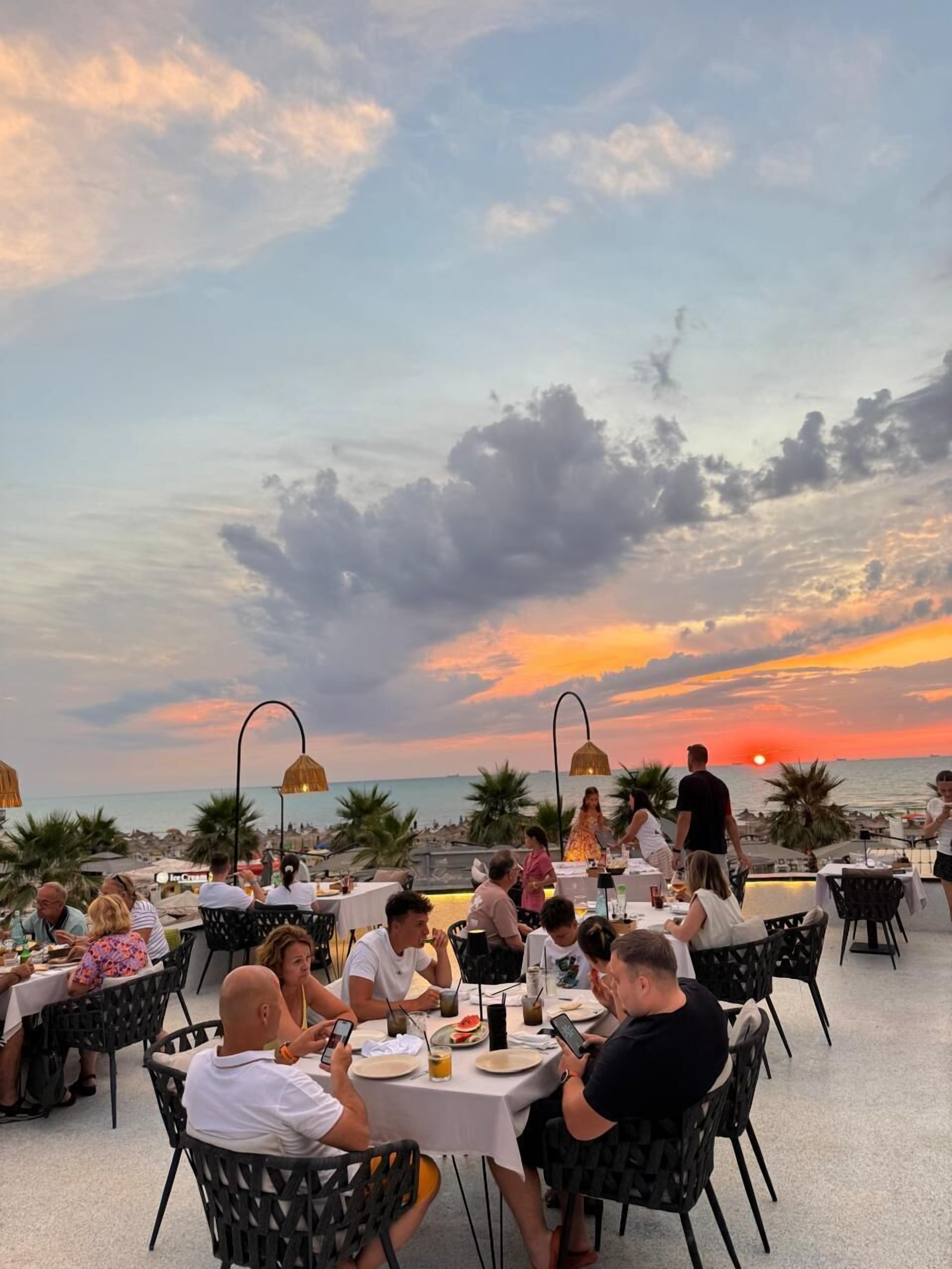 Un restaurant doté d’une terrasse offrant une vue sur la mer, avec des tables et des chaises rondes, et d’où l’on peut admirer le coucher du soleil.