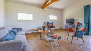 Un salon spacieux avec un plafond haut, des poutres en bois, un canapé gris, des fauteuils colorés, une petite table avec une plante et un meuble pour la télévision.