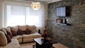 Ein Wohnzimmer mit einer Couch, einem Couchtisch, einem an einer Ziegelwand montierten Fernseher und einem großen Fenster mit Jalousien.