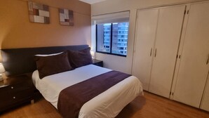 Ein Schlafzimmer mit einem Bett, einem Nachttisch, einem Kleiderschrank und einem Fenster mit Blick auf Gebäude.