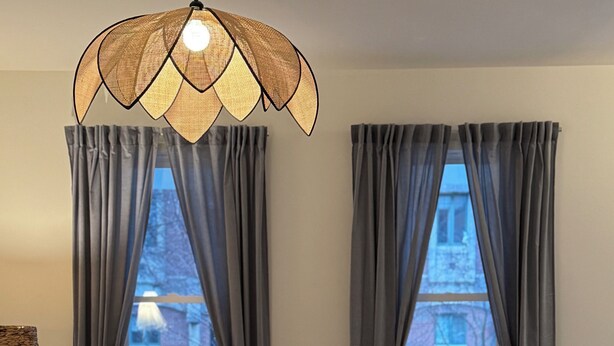 Ein Wohnzimmer mit einer Couch, einem runden Couchtisch und einer Lampe. Zwei Fenster mit Vorhängen und ein Blick auf Gebäude im Freien.