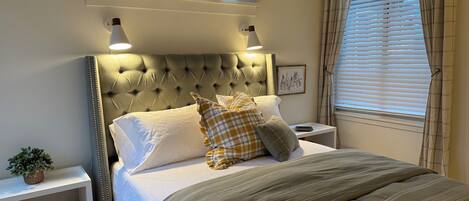 A neatly made bed with a tufted headboard, a bedside table with a lamp, a small plant, and a window with blinds.