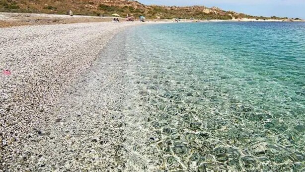 Una playa de guijarros con agua cristalina, una costa lejana y un paisaje montañoso al fondo.