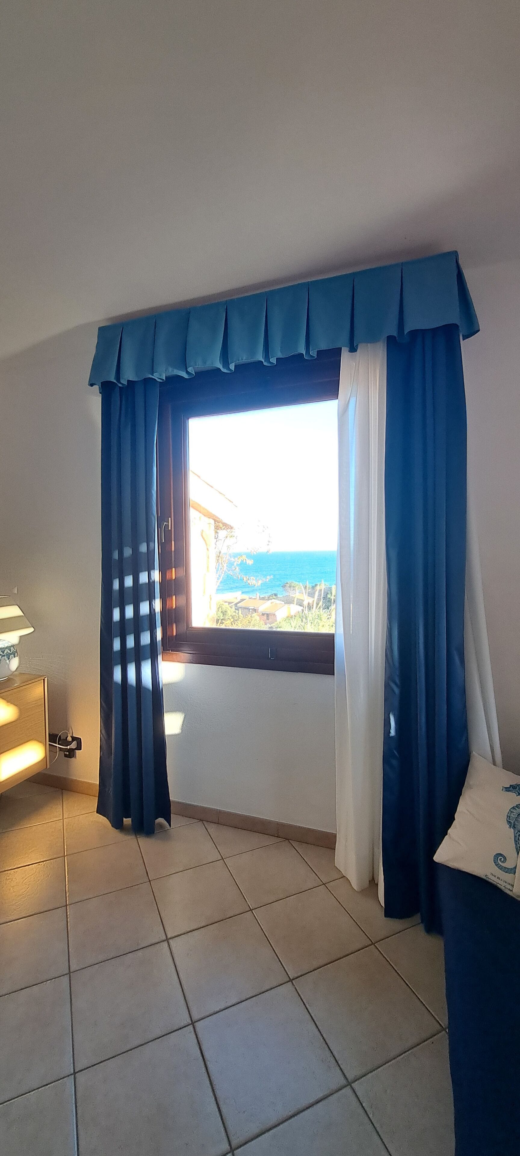 Een kamer met uitzicht op zee, een raam met blauwe gordijnen, een lamp en een betegelde vloer.