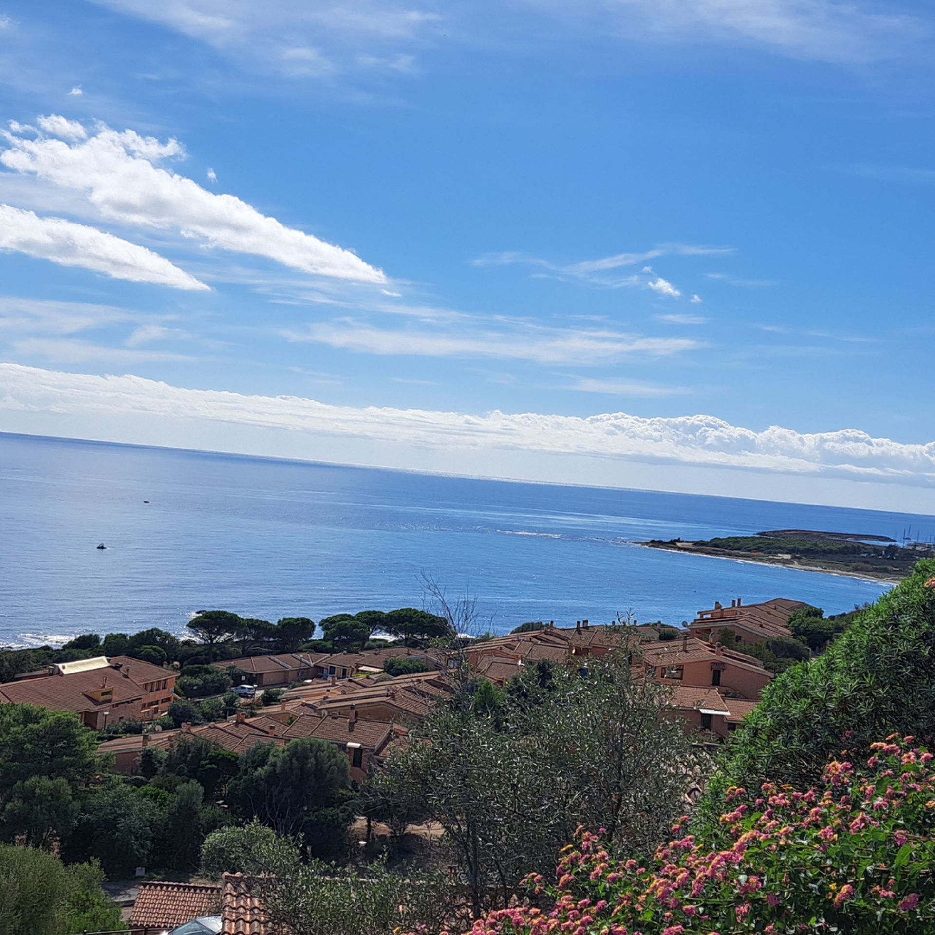 A coastal village with terracotta-roofed buildings, a clear blue sky, and the sea in the background.