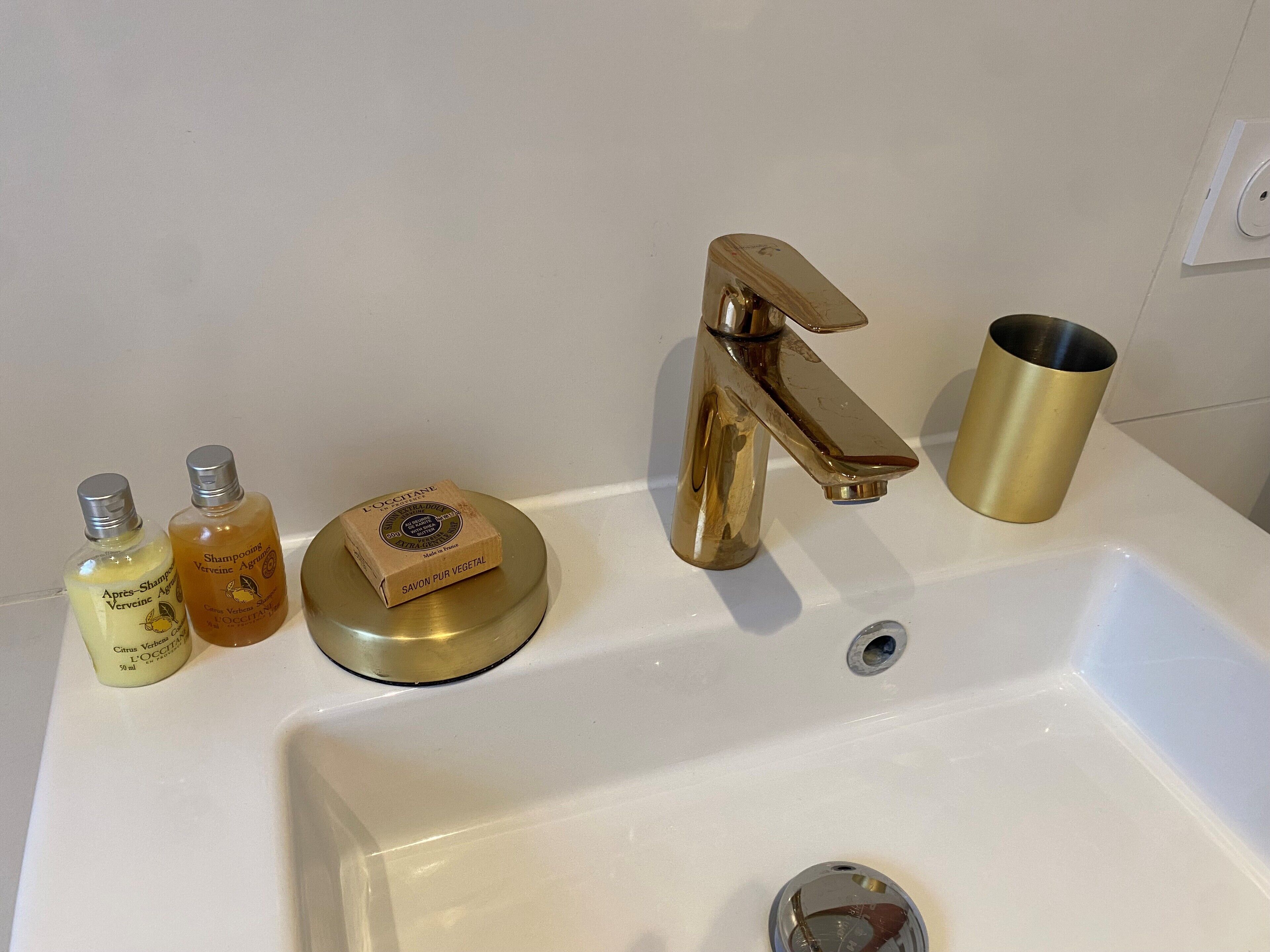 A bathroom sink with two bottles of shampoo, a soap dish, a gold-colored faucet, and a gold-colored cup.