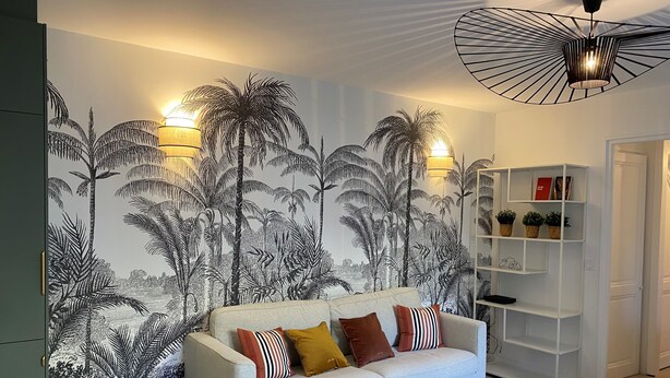 Una sala de estar con un sofá de gris claro, pared con diseño y una estantería blanca.
