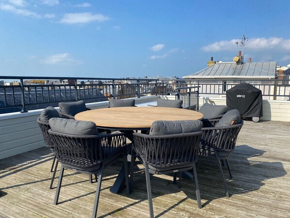 A round wooden table with six chairs, a grill, and a view of the city skyline.