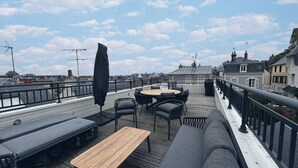 Una terraza en la azotea con mobiliario exterior, incluyendo una mesa redonda y sillas, además de una mesa de madera.