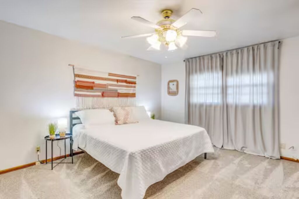 Quarto com cama, ventilador de teto, janela com cortinas e uma obra de arte na parede.