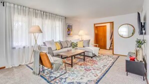 Una sala de estar moderna con un conjunto de sofás, una mesa de centro y un tapete estampado. Hay una lámpara, un espejo y una puerta que da a otra habitación.