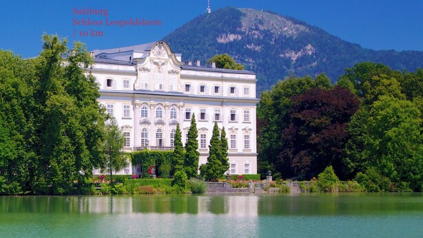 A large, classical building with a symmetrical facade, surrounded by lush greenery and a calm lake, with a mountain in the background. - Quartier No.7 - Serviced living at the gates of Salzburg (Grödig)