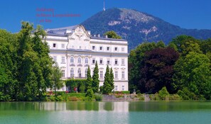 Un edificio grande de estilo clásico con una fachada simétrica, rodeado de vegetación exuberante y un lago tranquilo, con una montaña al fondo.
