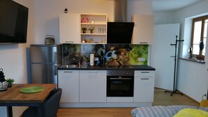 A compact kitchen with a built-in oven, a microwave, and a sink. There is a dining table with chairs, a bed with a striped blanket, and a window with blinds. - Quartier No.7 - Serviced living at the gates of Salzburg (Grödig)