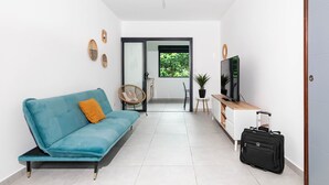 Een moderne woonkamer met een blauwe bank, een rieten stoel, een televisie, een bijzettafeltje en een koffer.
