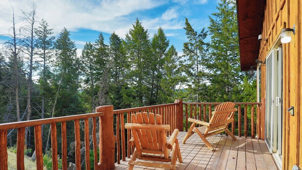 A wooden deck with two Adirondack chairs, a railing, and a view of a forest.