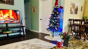 A living room with a television displaying a fireplace scene, a small decorated Christmas tree, and a coffee table with a red mug. - Charming Cozy Cabin in a PEACFUL MOUNTAIN setting w HOTTUB! Close to PigeonForge (Seymour)