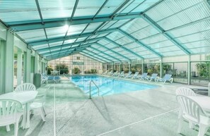 Une piscine couverte, dotée de chaises longues et de tables, entourée de murs en verre et surmontée d’un toit en métal.