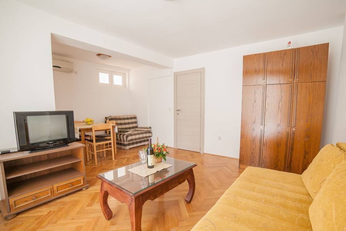 A living room with a wooden floor, a sofa, a coffee table, a television, a dining table with chairs, and a wardrobe.