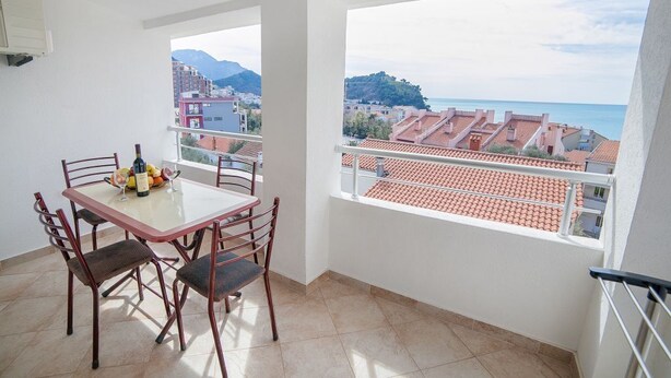 A balcony with a table and chairs, a view of the sea and buildings, and a tiled floor. - Ivan Apartmani (Petrovac)