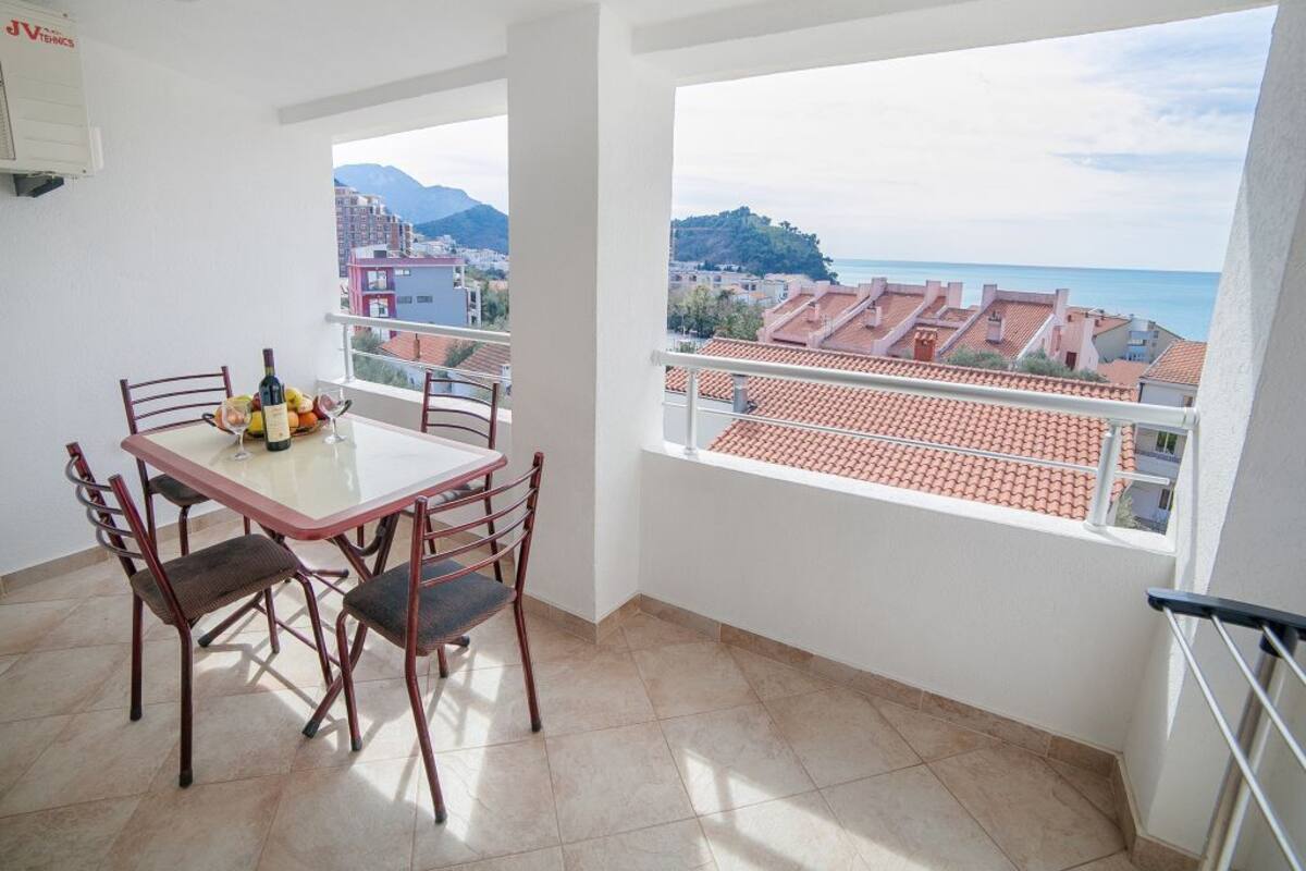 A balcony with a table and chairs, a view of the sea and buildings, and a tiled floor.