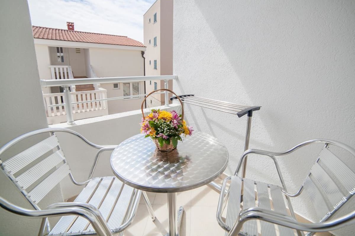 A balcony with a round glass table and two metal chairs, a wicker basket with flowers, and a view of buildings in the background.