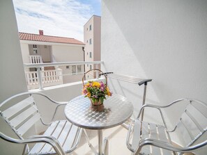A balcony with a round glass table and two metal chairs, a wicker basket with flowers, and a view of buildings in the background. - Ivan Apartmani (Petrovac)