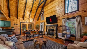 L’intérieur chaleureux d’un chalet avec des murs en bois, une cheminée en pierre, un coin repas et un téléviseur fixé au mur.