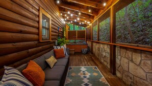 L’intérieur d’une cabane en rondins, avec un plafond en bois, un mur en pierre, un canapé avec des coussins et une vue sur la forêt à travers de grandes fenêtres.