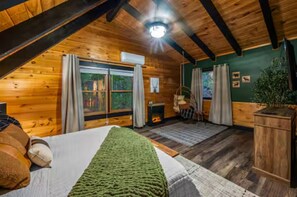 A cozy attic bedroom with wooden walls, a large bed, a fireplace, and a view of the outdoors. - Prime Location! 1mi to Parkway, Sauna, Game Room! (Pigeon Forge)