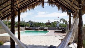 A hammock under a thatched roof, a pool, and lounge chairs on a sandy beach.