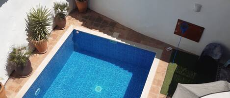 A swimming pool with tiled edges, surrounded by potted plants and a white wall with a tiled roof.