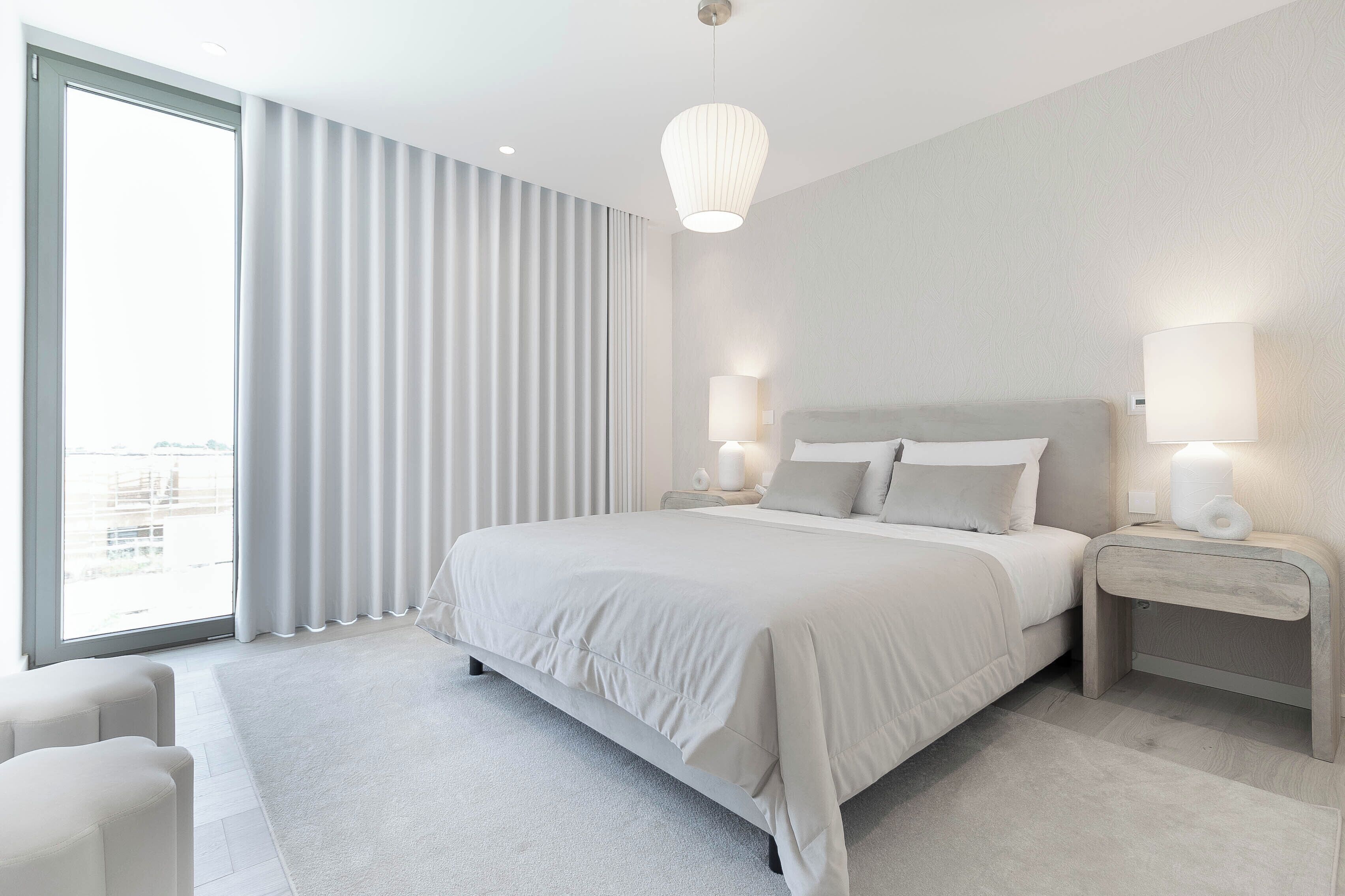 Una habitación de hotel moderna con una cama grande, dos mesitas de noche con lámparas, un ventanal con cortinas y vistas a la ciudad.